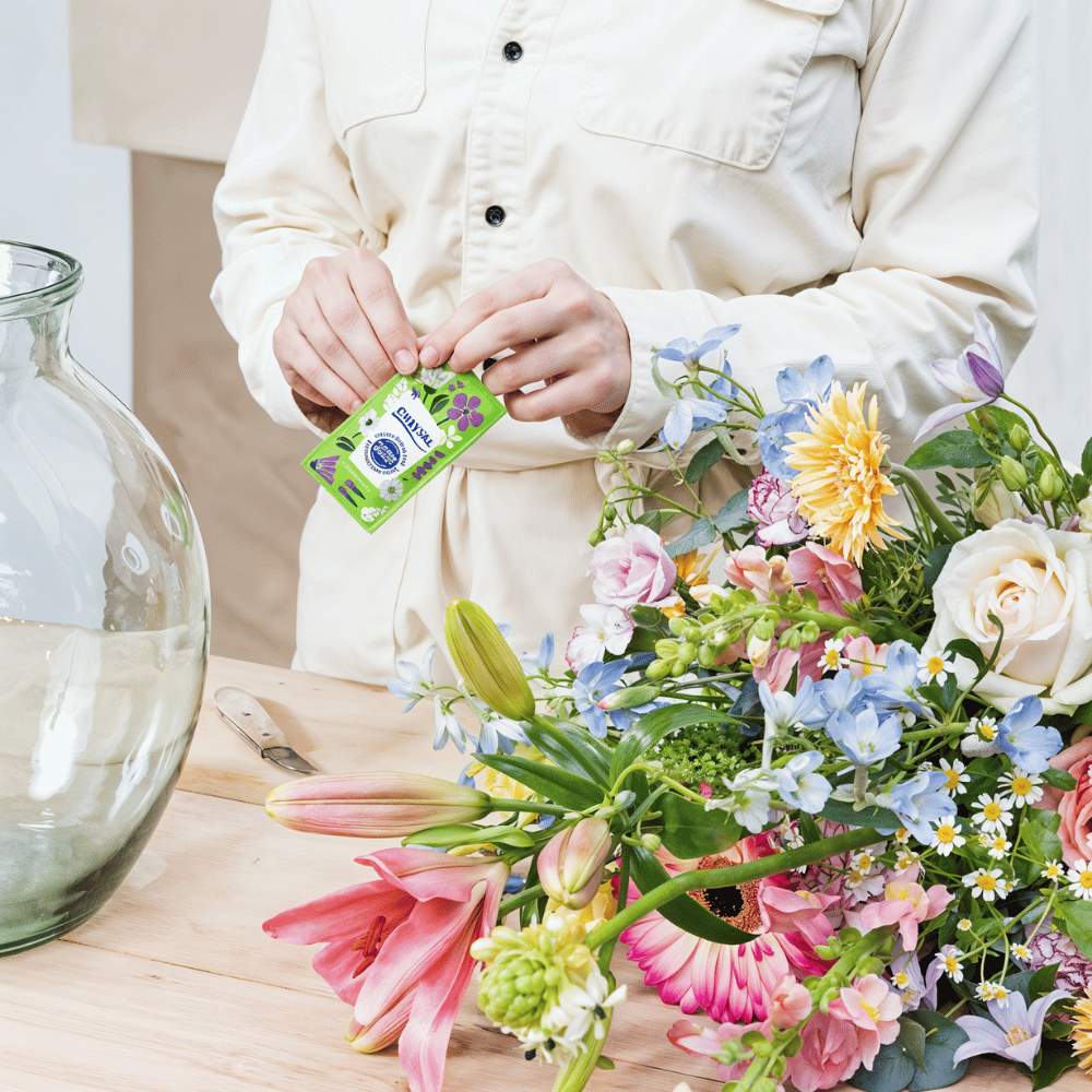 Cut Flowers Care: 5 Essential Tips to Keep Bouquets Fresh 6 Florist holding a packet of flower food beside a colorful flower bouquet and a glass vase.